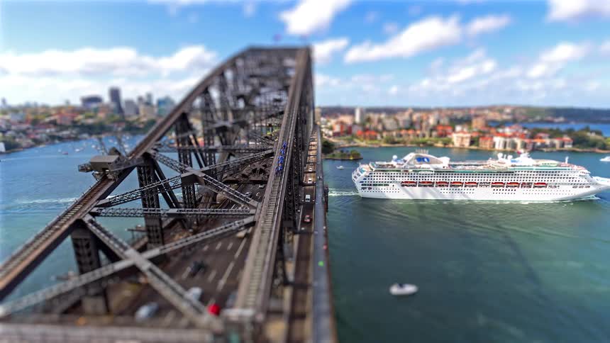 A tilt-shift time-lapse captures a massive cruise ship sailing under the iconic Sydney Harbour Bridge as traffic flows and other boats cruise the harbour