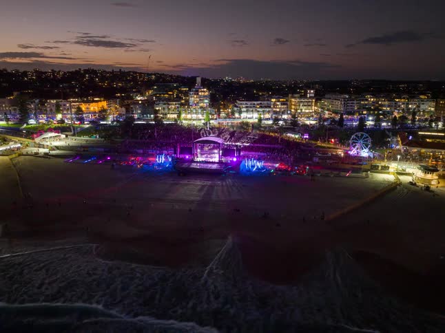 An aerial view shows a vibrant music festival lighting up a beach at dusk, with a large crowd gathered and a city twinkling in the background