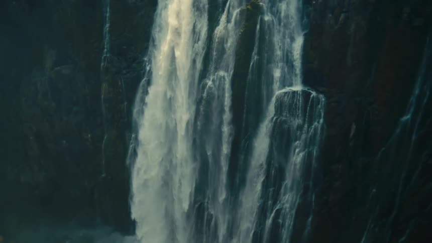 A powerful waterfall cascades over a massive cliff face, with the camera tilting down to follow the water's descent