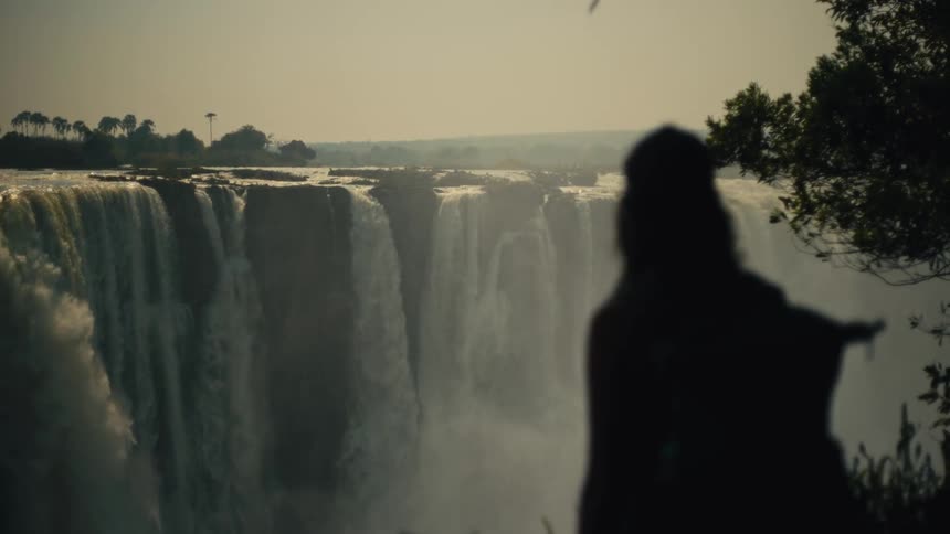 A silhouetted onlooker gazes out at the magnificent and powerful Victoria Falls in a serene, wide shot