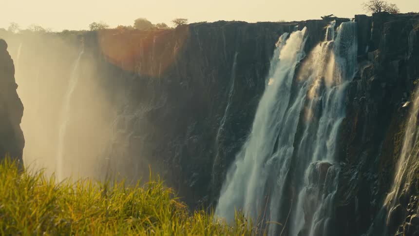 Golden sunlight illuminates the mist rising from a powerful waterfall as it cascades down a rocky canyon, with sun-kissed grass in the foreground