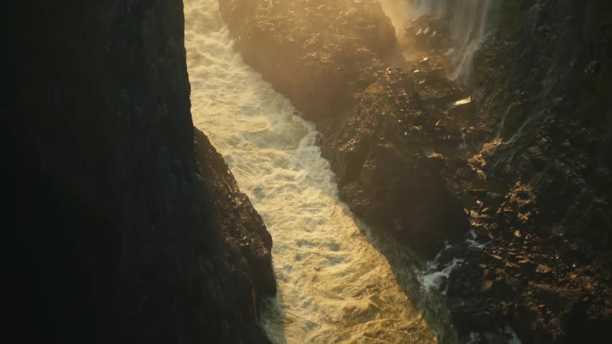 Golden hour light illuminates a raging river as it carves its way through a dark, rocky gorge