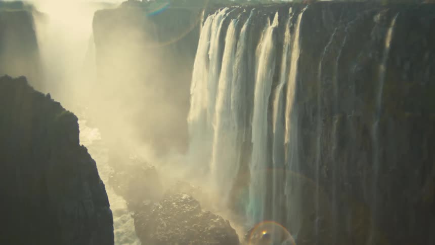 Sunlight shines through the mist of a powerful waterfall as it cascades into a deep, rocky canyon, with the camera tilting upwards to reveal its full scale