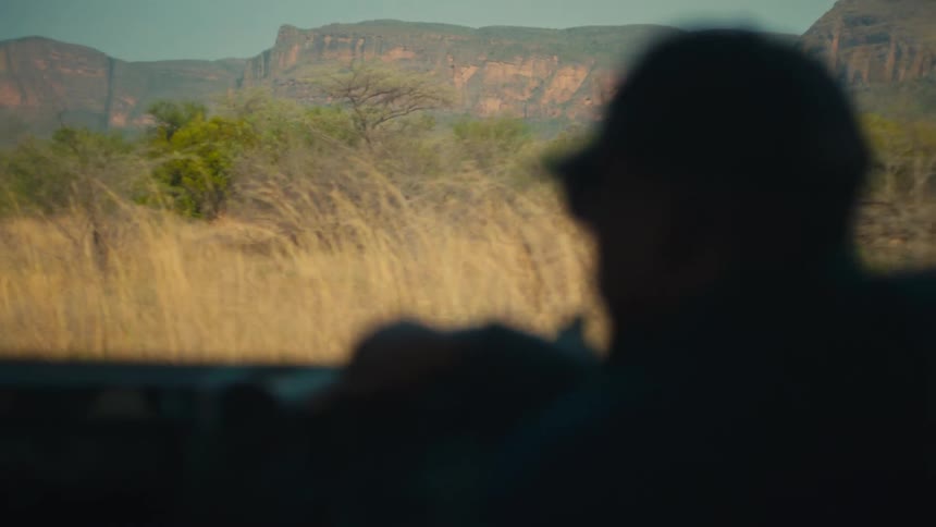 An over-the-shoulder perspective of a safari guide navigating a rugged jeep through the African savanna with distant mountains on the horizon