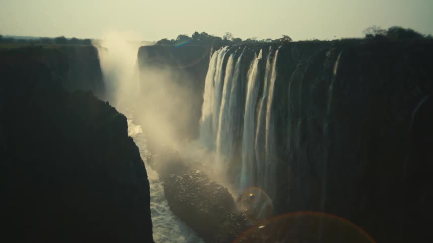 A stunning wide shot of a massive waterfall plunging into a deep, misty gorge, backlit by the warm glow of the sun