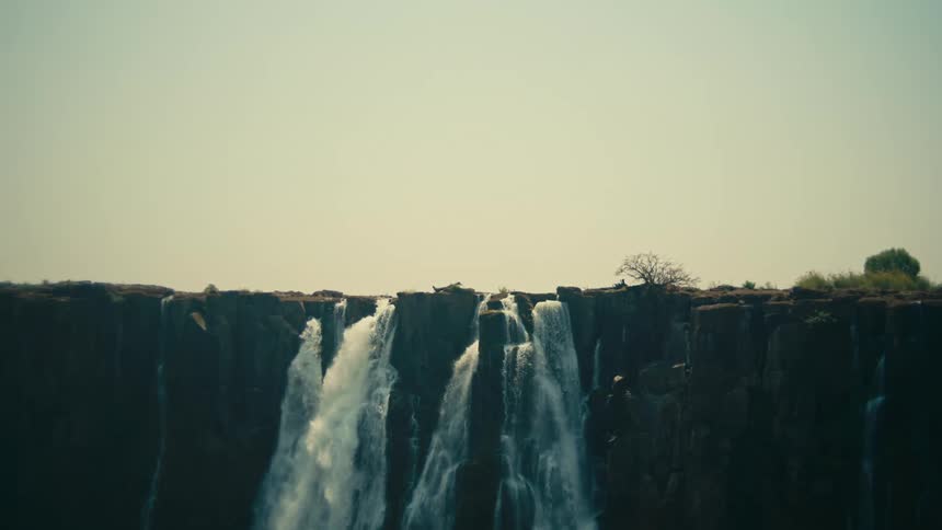 A powerful waterfall crashes over the edge of a wide, rocky cliff under a hazy sky
