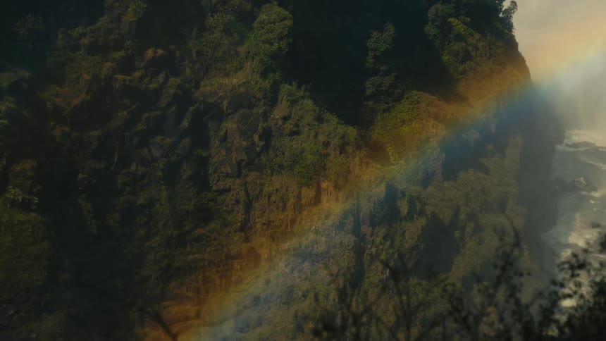 A stunning rainbow shines through the mist over a lush, tree-covered canyon