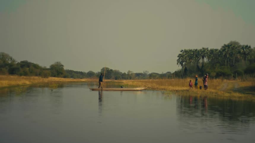 A man poles a traditional mokoro boat across a calm river while his family waits on the bank