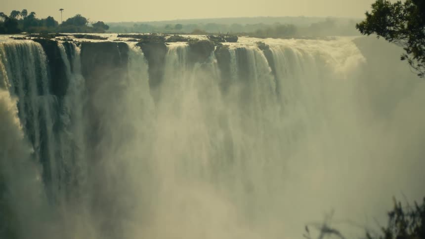 A powerful waterfall thunders over the edge of a vast cliff, creating a thick cloud of mist in the hazy afternoon light