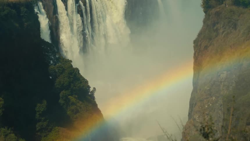 A vibrant rainbow arcs across the misty gorge of a powerful, cascading waterfall