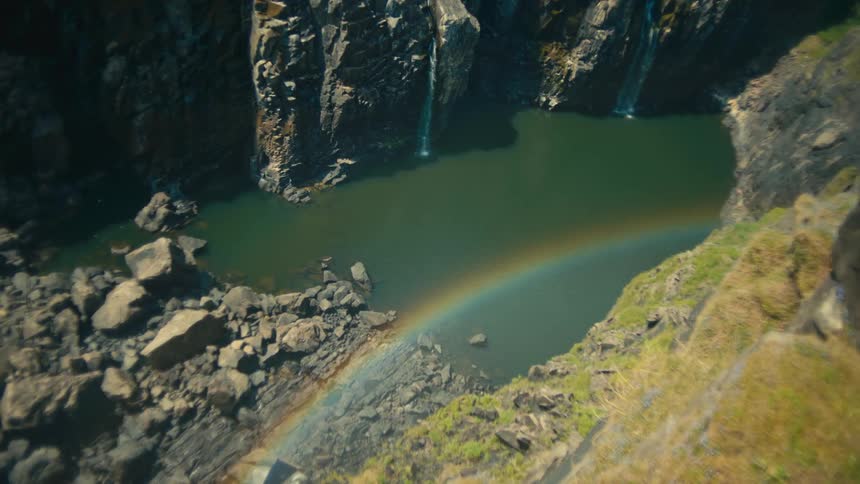 A vibrant rainbow arcs over a tranquil pool at the base of waterfalls in a majestic, rocky canyon