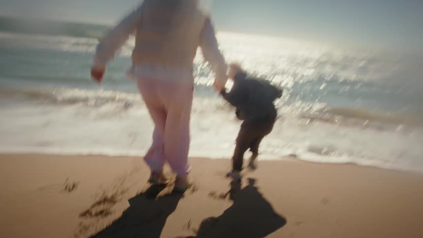 In a dreamy slow-motion shot, a mother and her young son joyfully run hand-in-hand along the sandy beach toward the sunlit ocean waves