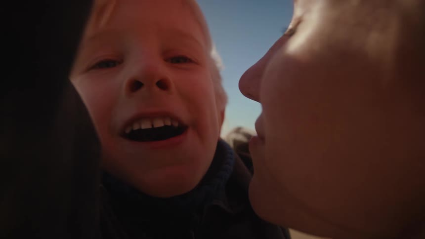 An intimate, sun-flared close-up shows a mother kissing her smiling son on a sandy beach, capturing a moment of pure joy in slow motion