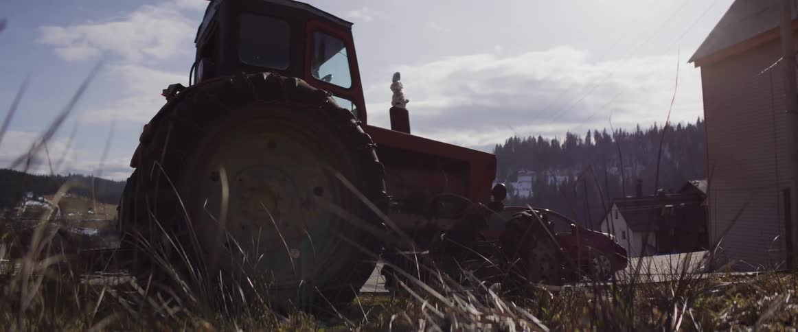 A low-angle view shows a vintage red tractor parked by a rural road with dry grass swaying in the foreground