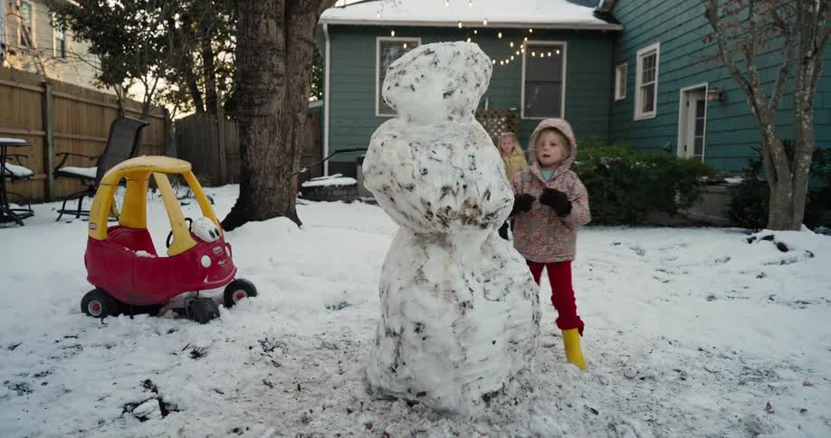 A toddler wearing a hooded coat and bright yellow boots adds finishing touches to a lumpy snowman in a snowy backyard