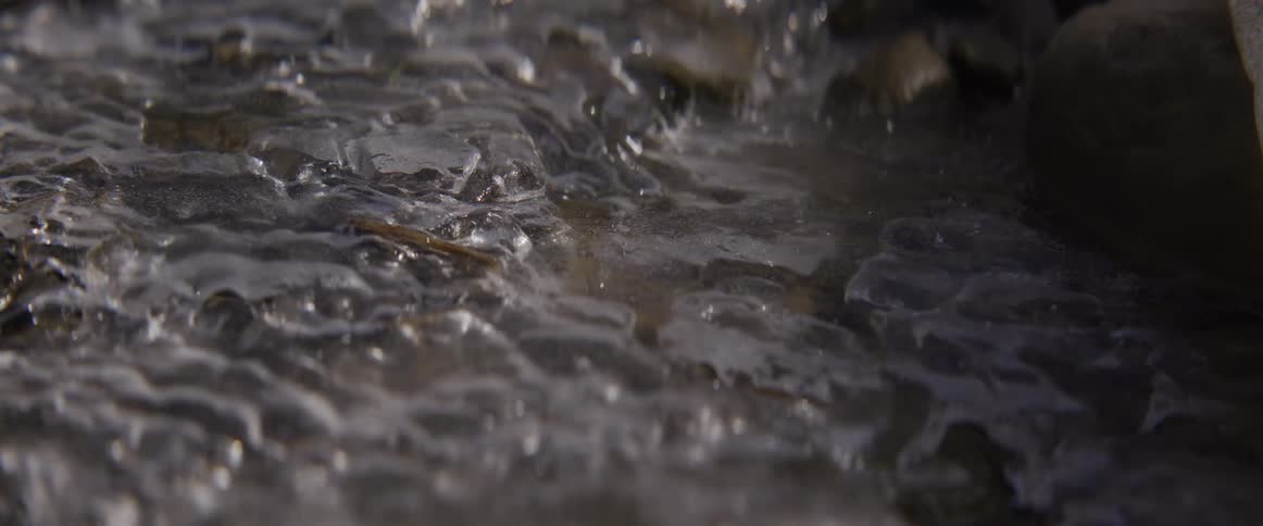 An intimate slow-motion close-up of water flowing over a partially frozen stream
