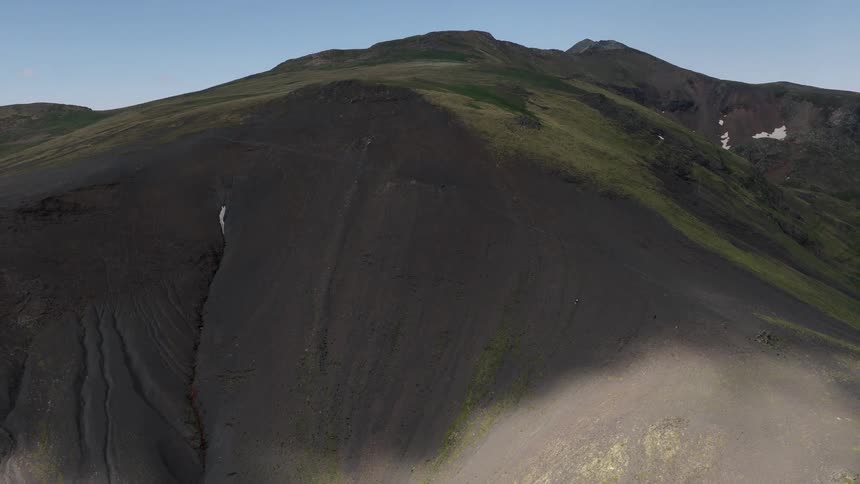 A tiny hiker traverses the immense, dark, and grassy slopes of a mountain in this epic aerial pullback shot