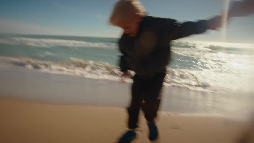 A mother and her young son energetically run and play together, holding hands along the sunny ocean shore