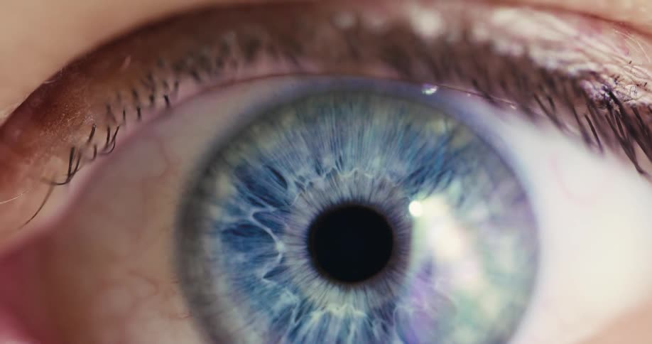 A human eye is revealed from darkness in a macro shot, its blue iris reflecting colorful light as the pupil adjusts