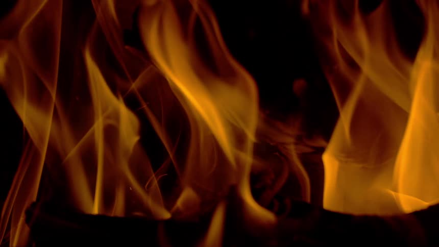 A close-up slow-motion shot of bright orange and yellow flames dancing over burning logs against a black background