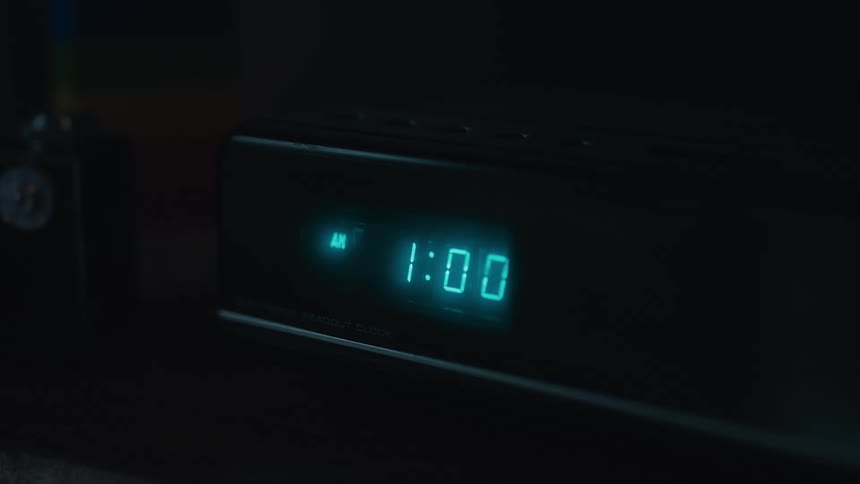 A digital alarm clock in a dark room changes from 12:59 AM to 1:00 AM