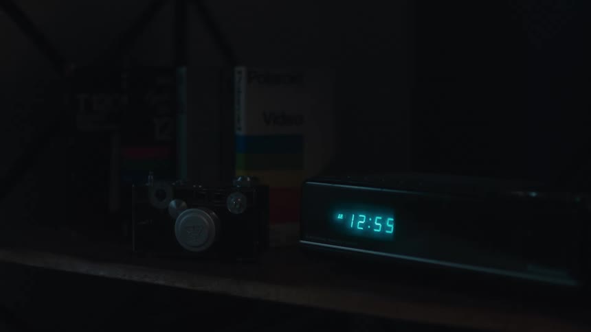 A glowing digital alarm clock on a shelf displays the time next to a vintage camera in a dark room