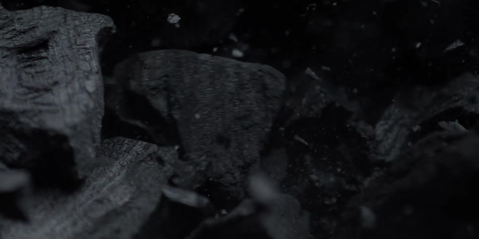 A macro shot captures charcoal chunks and dust falling in super slow motion