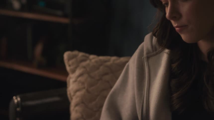 A young woman in a comfortable hoodie reads a book intently in a dimly lit, cozy room at home