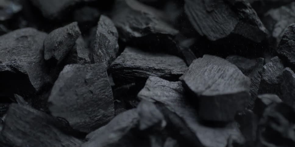 A dramatic slow-motion macro shot captures the intricate textures of black charcoal pieces as dust particles float and settle