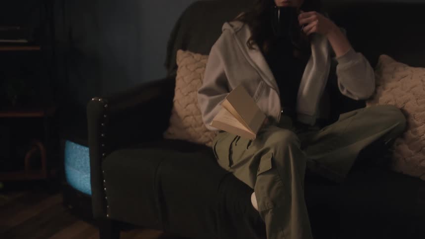 A young woman relaxes on a cozy couch in a dimly lit room, reading a book and sipping a warm beverage from a mug