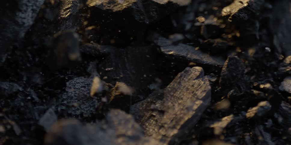 A dramatic slow-motion macro shot panning over a pile of dark charcoal pieces with warm highlights