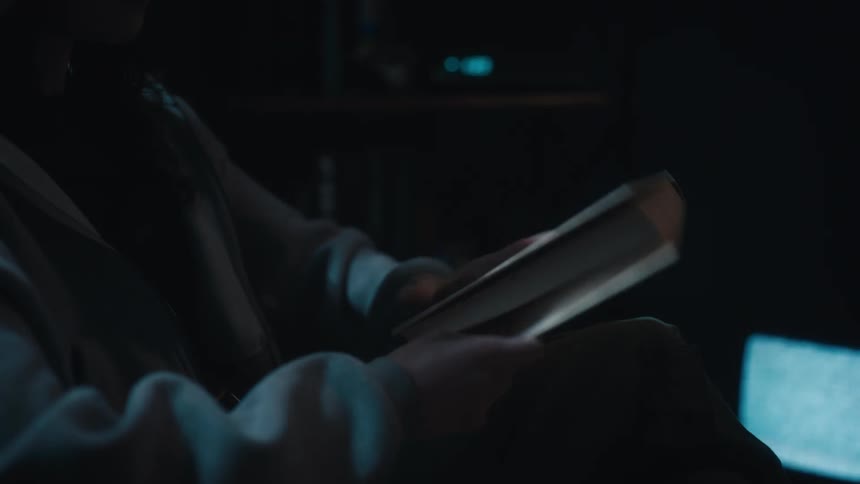 A woman reading a book in a dark room suddenly realizes the late hour and quickly gets up
