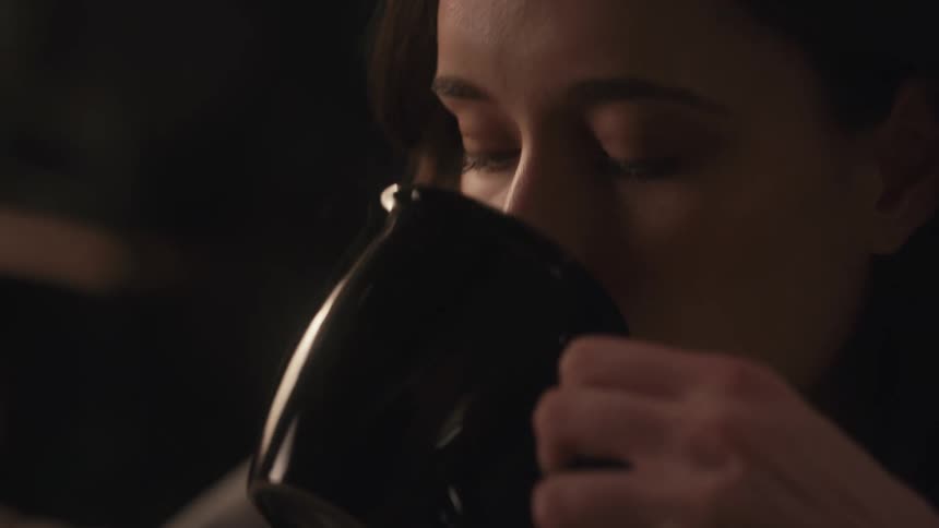 An intimate, slow-motion close-up of a contemplative woman sipping a warm beverage from a black mug in a dimly lit room