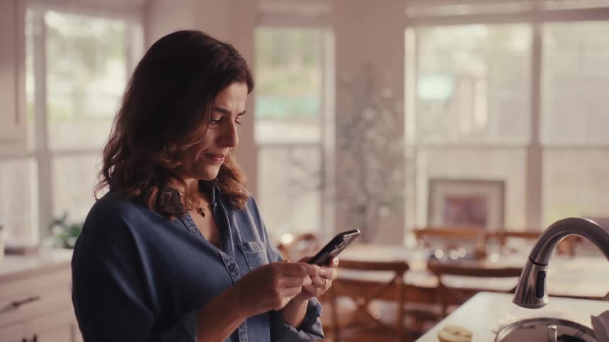 A worried woman stands in her kitchen, checks her phone, smiles, and takes a breath of relief