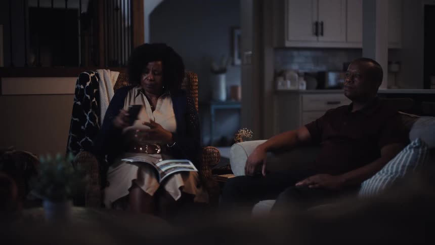 An older African American couple relax in their living room at night; the man watches TV while the woman reads a magazine and checks her phone