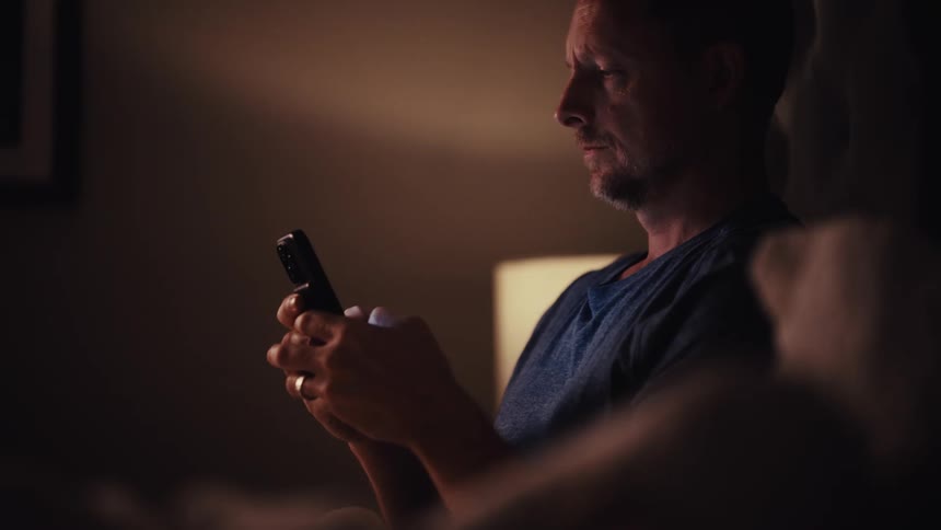 A man reading a spiritual book in a dark room gets distracted by his smartphone