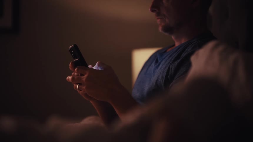 A man reading a spiritual book in bed at night gets distracted and starts scrolling on his smartphone