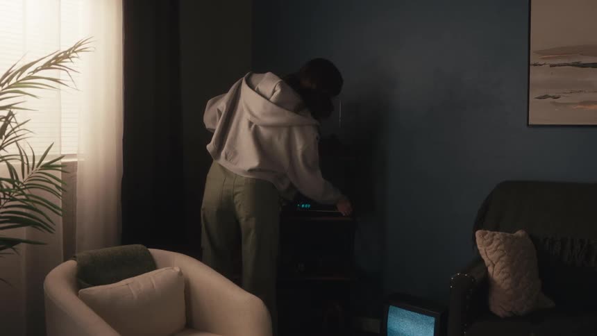 A young woman walks through her retro living room, puts a hidden VHS tape into the VCR, and sits down on the couch to read a book