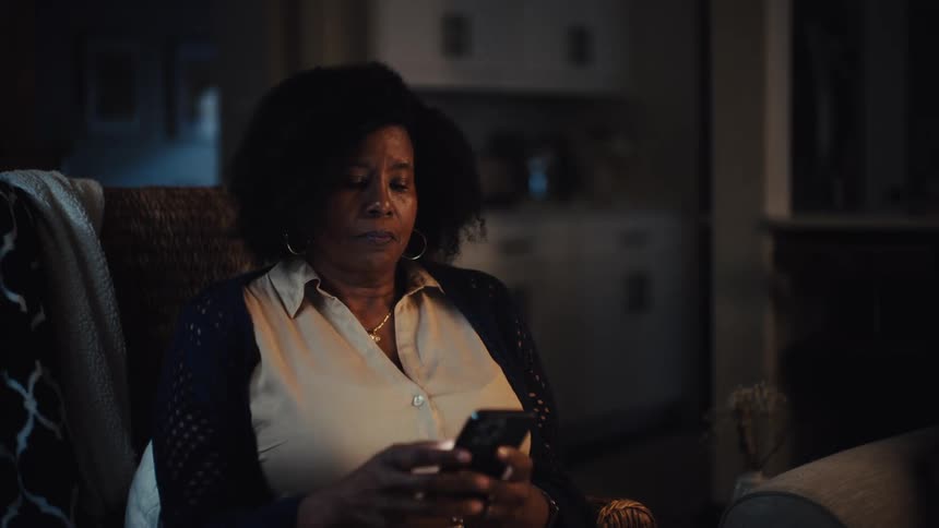 A thoughtful older woman sits alone in her dimly lit living room at night, browsing a magazine before picking up her phone