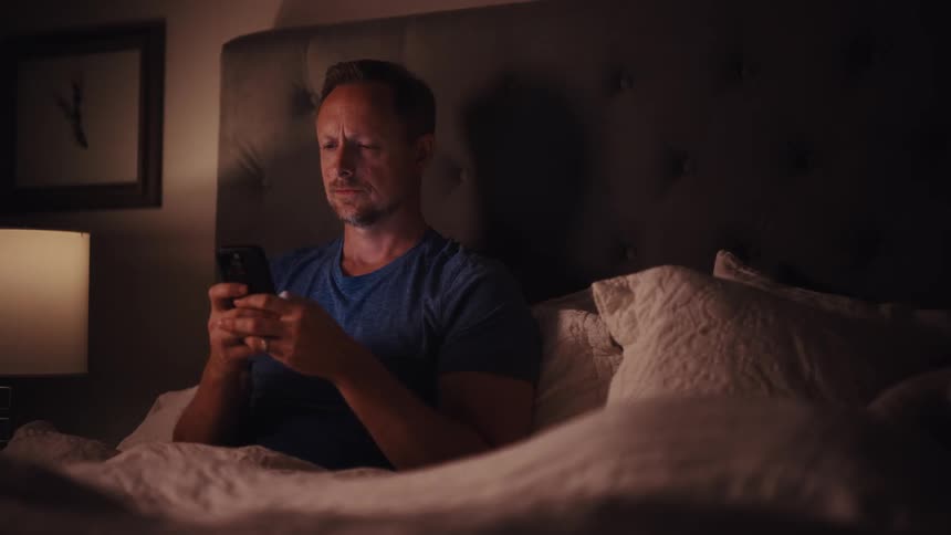 A man reading a spiritual book in bed at night puts it down to start scrolling on his smartphone instead