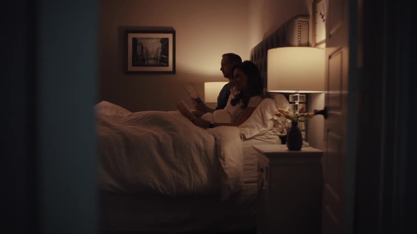 A couple winds down for the night in bed, with the woman reading a book before turning off her lamp to sleep