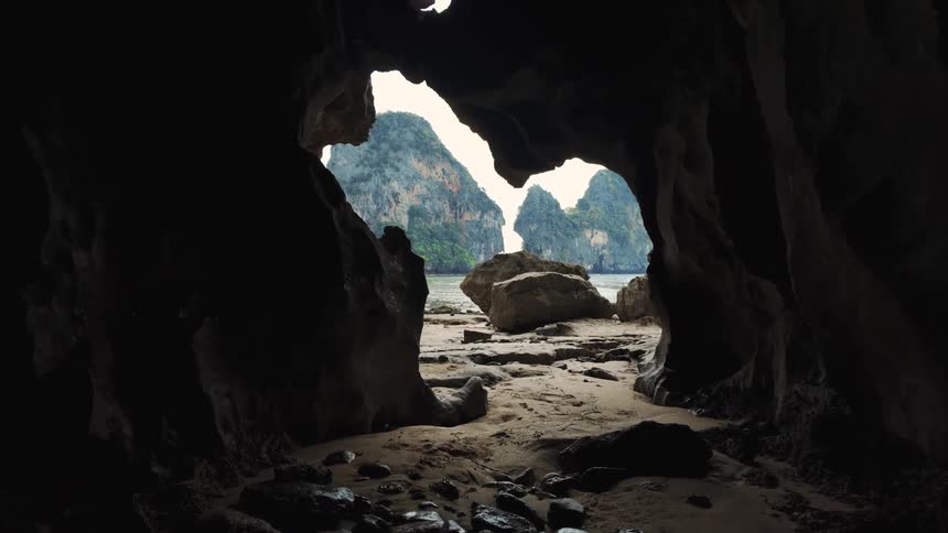 A slow zoom out from inside a dark coastal cave reveals a stunning view of a tropical beach and limestone islands
