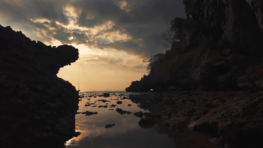 A sailboat glides across a tranquil ocean cove during a dramatic sunset, framed by silhouetted rock formations
