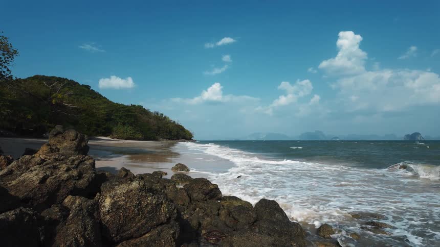 A slow-motion view of gentle ocean waves washing ashore on a secluded, rocky tropical beach under a bright blue sky