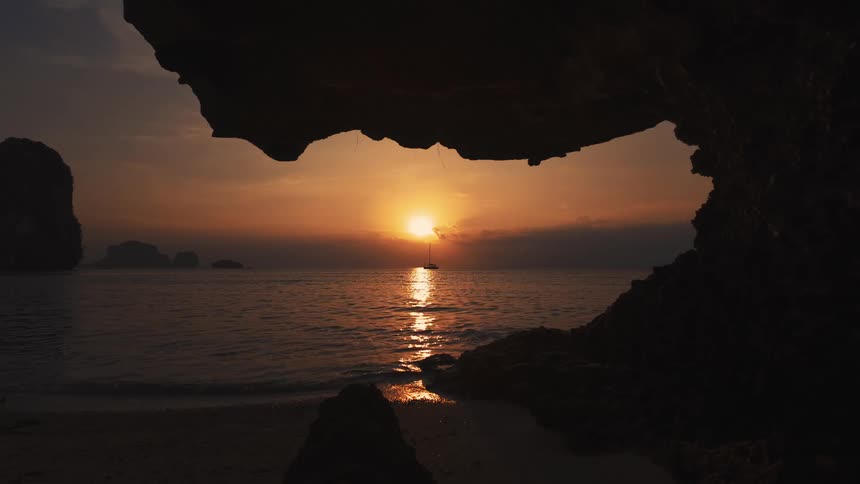 A picturesque sunset over the sea is framed by a rocky cave silhouette with a sailboat on the horizon