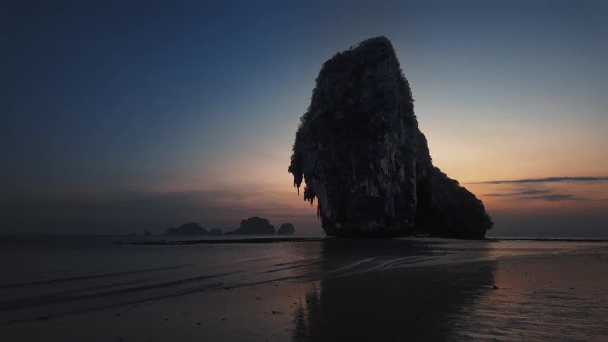 A majestic limestone karst is silhouetted against a colorful sunrise sky on a tranquil beach at low tide