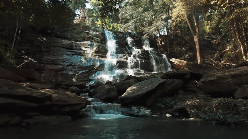 A beautiful multi-tiered waterfall cascades down a rocky cliff into a tranquil pool in a lush, sun-drenched forest