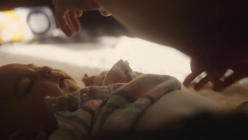 A tender, slow-motion close up shows a parent's hands gently dressing their newborn baby in a softly lit room
