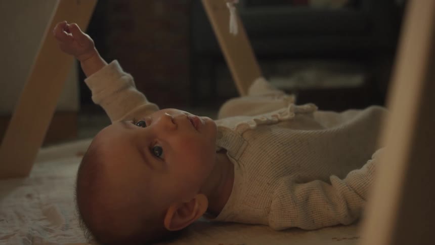 A close-up of a sweet baby in a white onesie yawning and stretching while lying under a wooden play gym in a cozy room