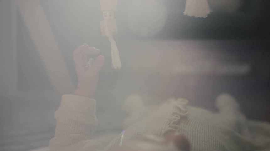 A newborn baby's tiny hand reaches out in slow motion to touch a hanging wooden toy mobile in soft, dreamy light